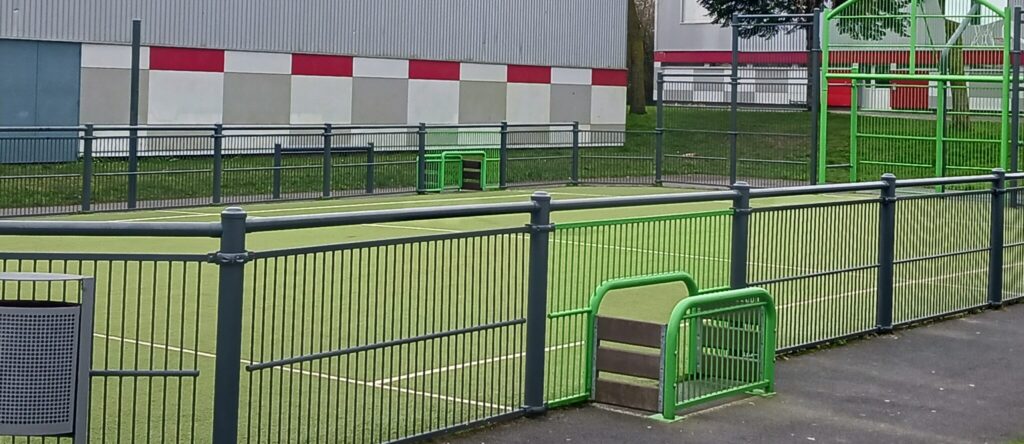 Terrain sportif extérieur, grillage vert, bâtiment industriel.