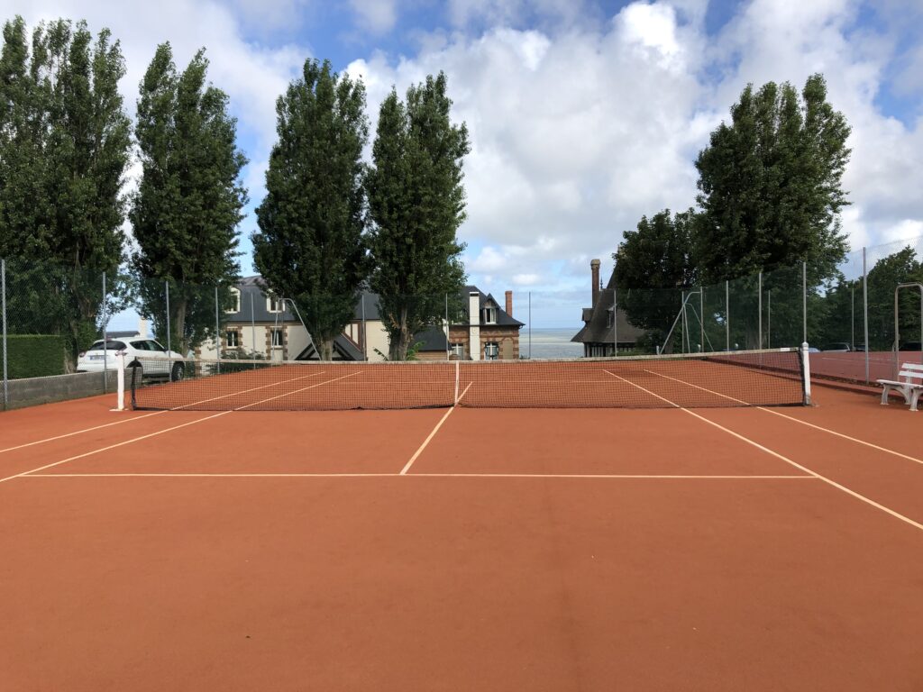 Court de tennis extérieur, terre battue, arbres, ciel nuageux.